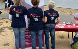 3 Mitarbeiter vom Hospiz stehen am See vor dem Hospiz-Banner. Die Mitarbeiter haben ein mit dem Hospiz-Logo bedrucktes T-Shirt an