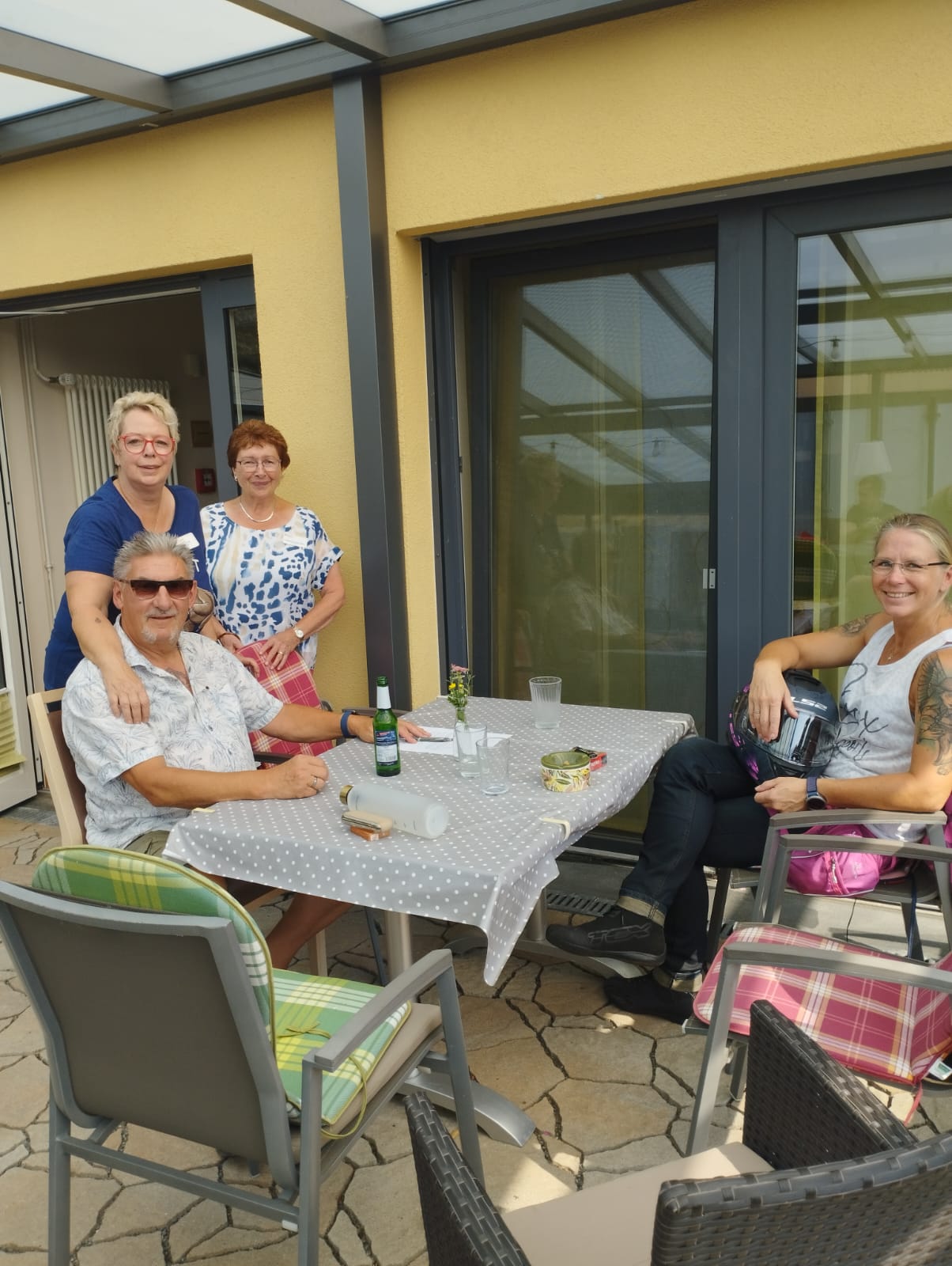 Ein Mann und eine Frau sitzen am Tisch, eine ehrenamtliche Mitarbeiterin und unsere Küchenfee stehen auf der Terrasse Ein Mann und eine Frau sitzen am Tisch, eine ehrenamtliche Mitarbeiterin und unsere Küchenfee stehen auf der Terrasse