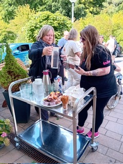 2 Frauen gießen Wasser in ein Glas und im Hintergrund stehen Menschen