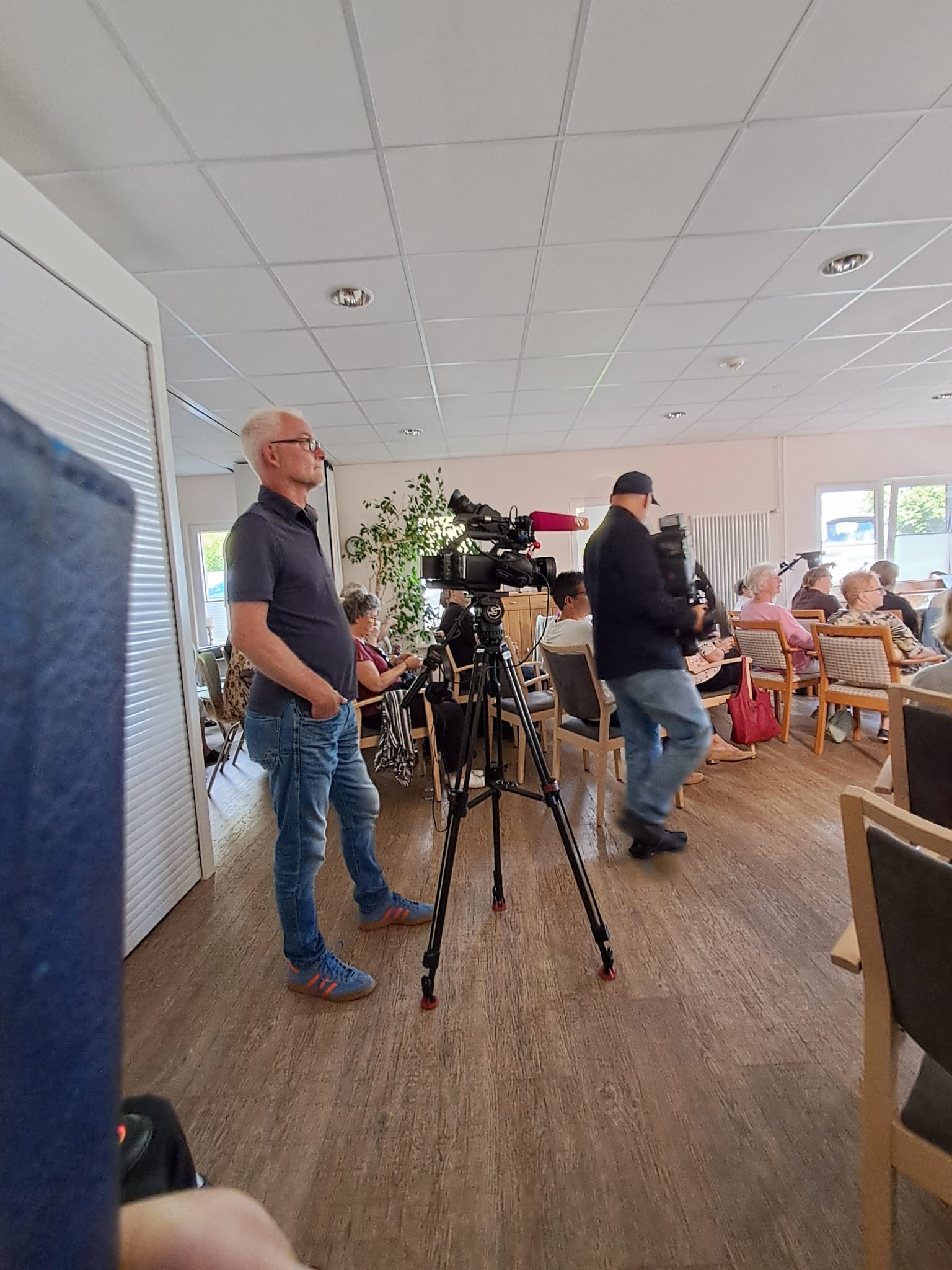 Mehrere Menschen sitzen im Veranstaltungsraum vom Hospiz. 2 Kameramänner sind ebenfalls anwesend