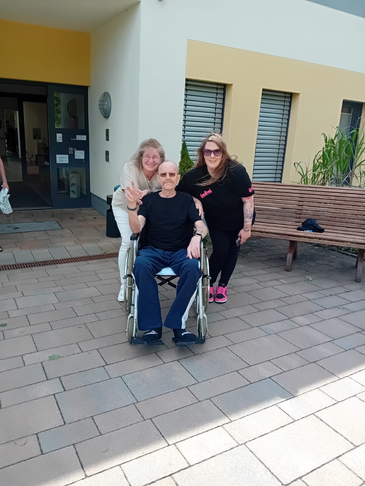 Ein Hospizgast sitzt in seinem Rollstuhl vor dem Hospizeingang. Hinter ihm stehen 2 Frauen