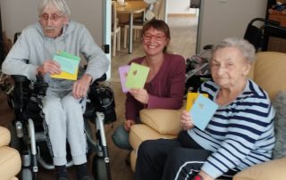 Mann im Rollstuhl, 2 Frauen halten sich im Hospiz-Wohnzimmer auf halten bunte Karten in den Händen