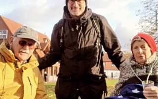 Ein Mann und eine Frau halten sich an den Händen. Ein anderer Mann hat jeweils einen Arm auf die Schulter des anderen Mannes und der Frau gelegt