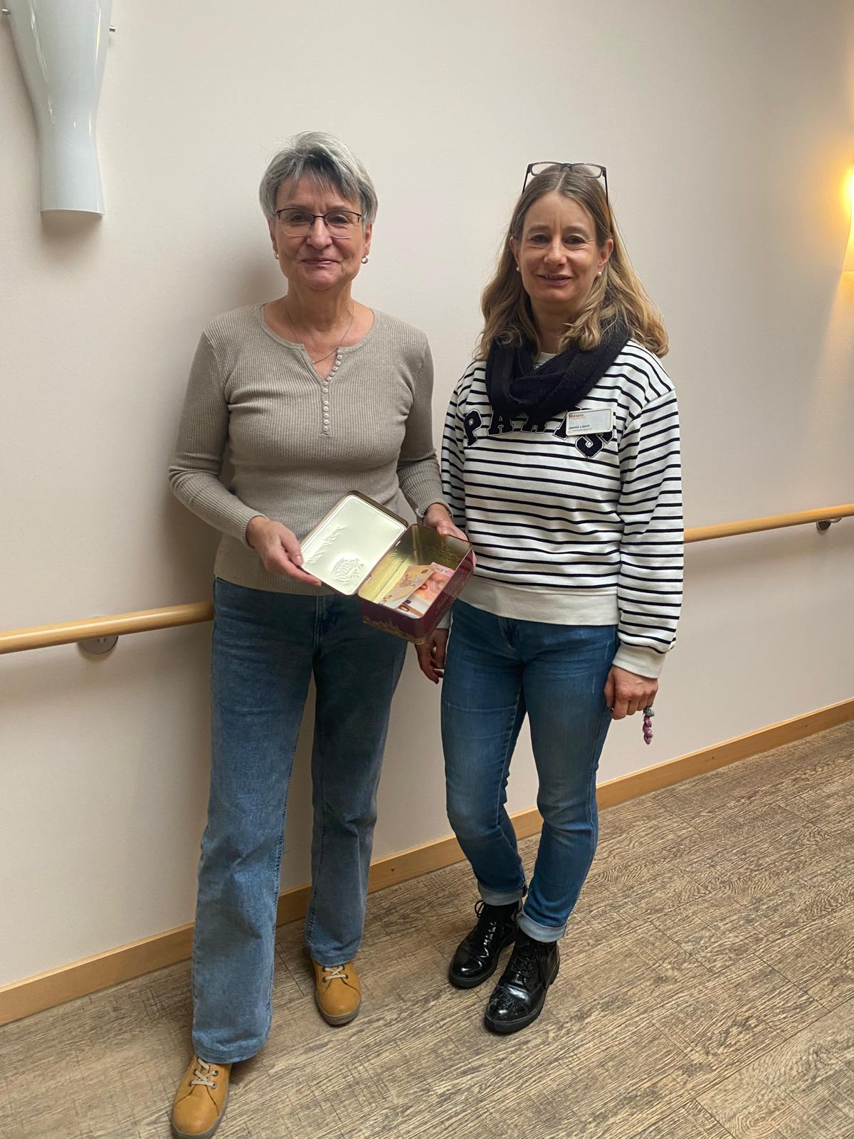 Zwei Frauen stehen vor einer Wand. Die eine Frau hält eine Schachtel mit Geldspenden in der Hand