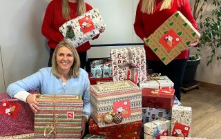 3 Frauen stehen um einen Wagen mit vielen Weihnachtsgeschenken, 2 Frauen davon haben Weihnachtsmützen auf