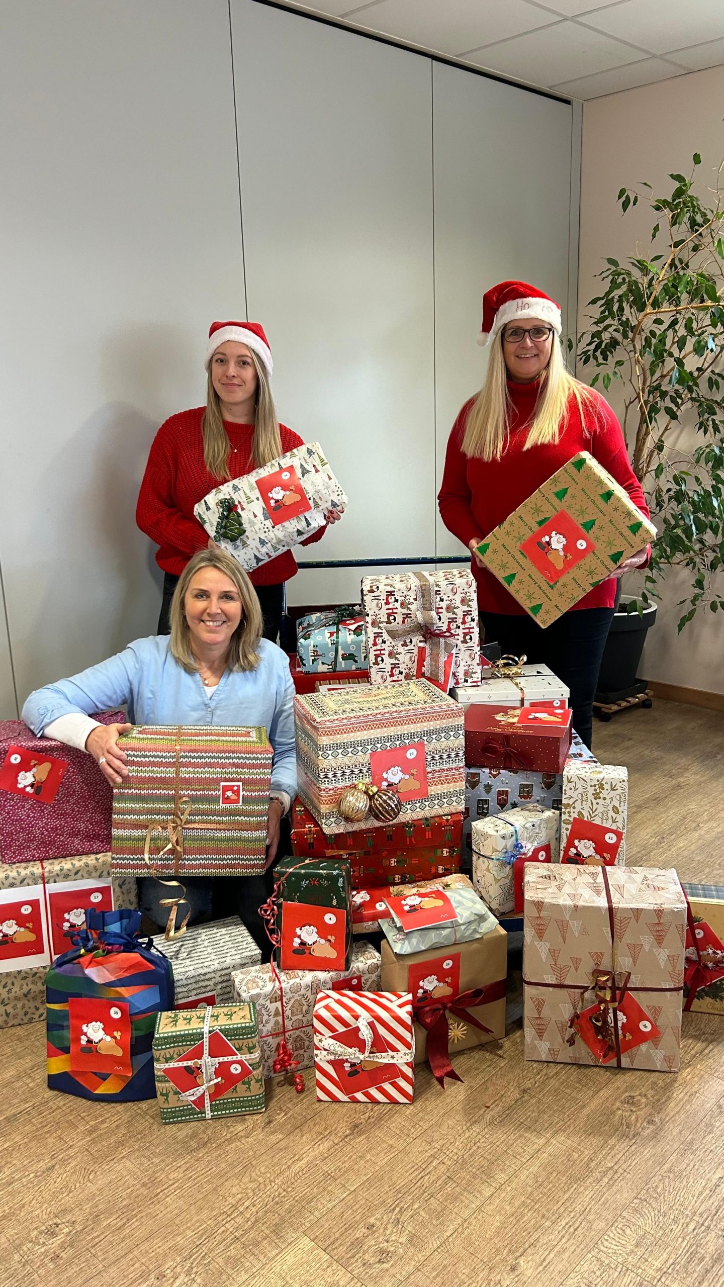 3 Frauen stehen um einen Wagen mit vielen Weihnachtsgeschenken, 2 Frauen davon haben Weihnachtsmützen auf