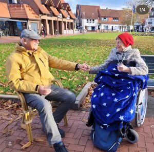 Ein Mann sitzt auf einer Bank. Eine Frau sitzt im Rollstuhl. Beide halten sich die Hände