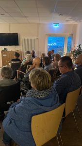 Mehrere Menschen sitzen auf Stühlen und hören den Musikern mit dem Akkordeon zu