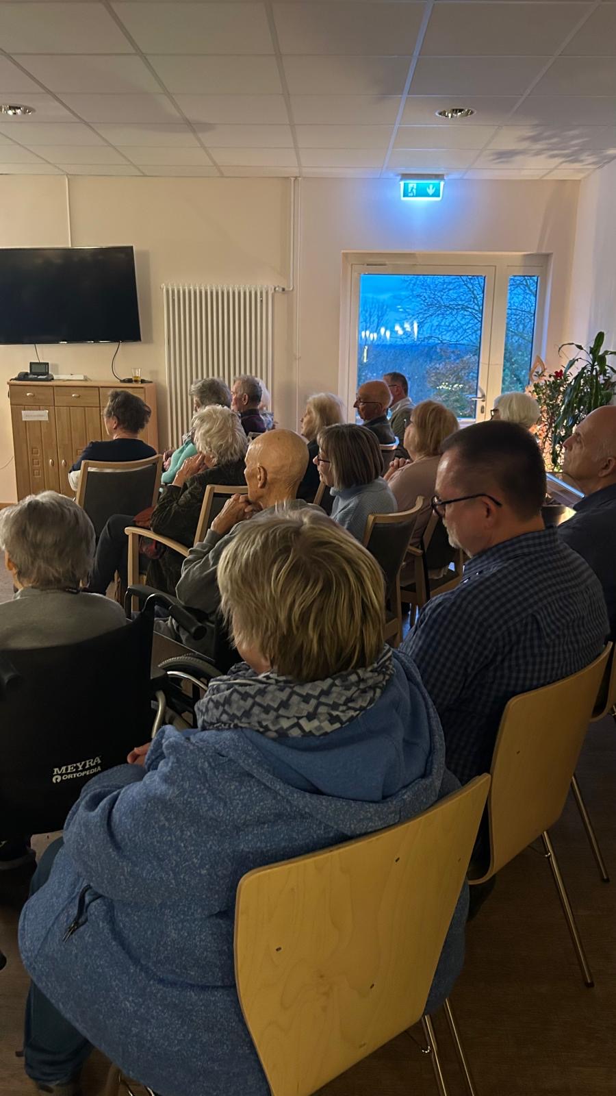 Mehrere Menschen sitzen auf Stühlen und hören den Musikern mit dem Akkordeon zu Mehrere Menschen sitzen auf Stühlen und hören den Musikern mit dem Akkordeon zu