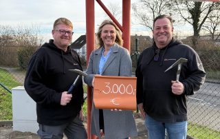 Zwei Männer und eine Frau stehen vor einem Firmenschild. Die Männer haben Werkzeug in der Hand, die Frau eine Ziegel. Auf der Ziegel steht die Spendensumme