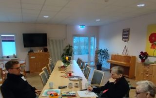 Ein Mann und zwei Frauen sitzen am Tisch. Auf dem Tisch liegen Blätter, Farbkästen, Pinsel und bunte Stifte