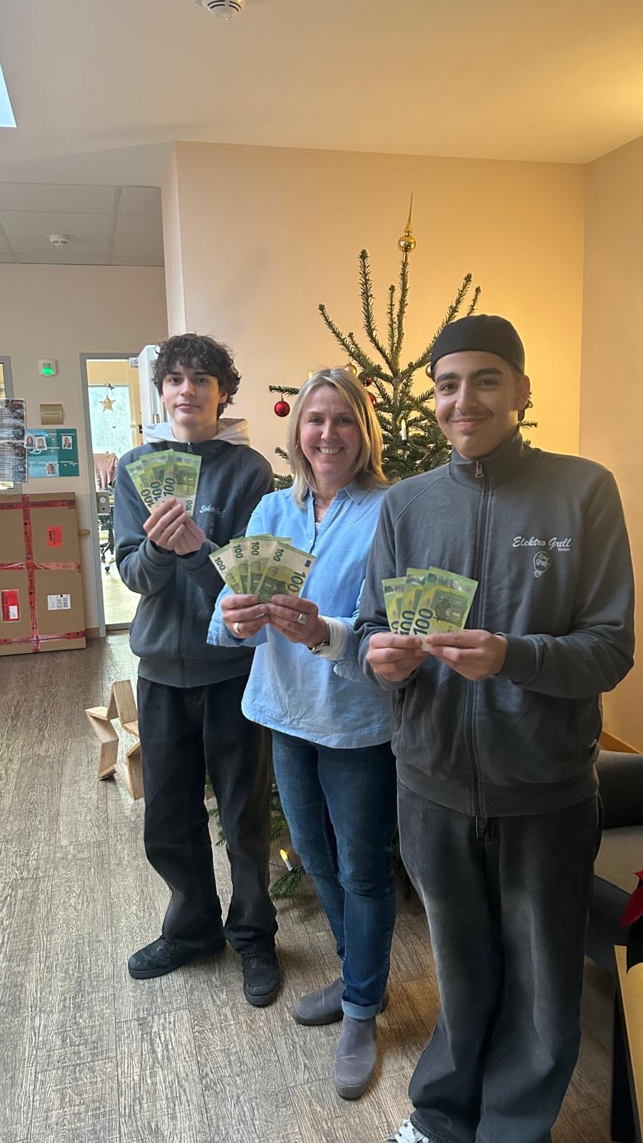 Eine Frau und zwei Jugendliche stehen vor einem Weihnachtsbaum und halten Geldscheine in den Händen
