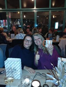 Zwei Frauen sitzen am Tisch, die eine Frau hält eine Tasse hoch und auf dem Tisch steht eine Weihnachtstüte