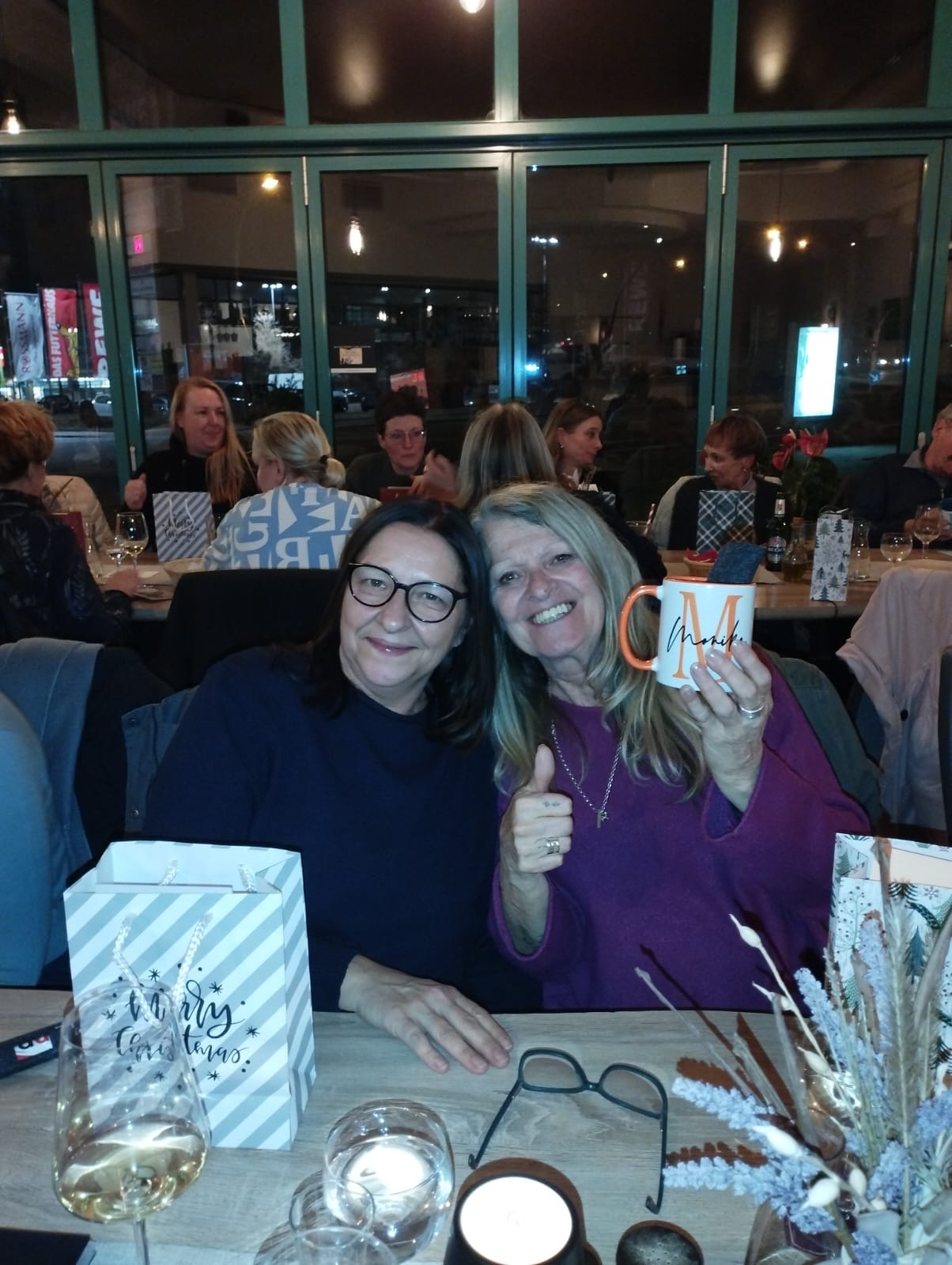 Zwei Frauen sitzen am Tisch, die eine Frau hält eine Tasse hoch und auf dem Tisch steht eine Weihnachtstüte Zwei Frauen sitzen am Tisch, die eine Frau hält eine Tasse hoch und auf dem Tisch steht eine Weihnachtstüte