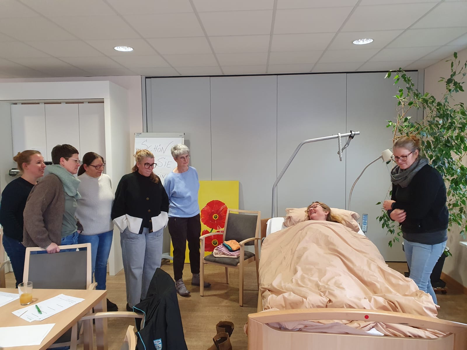 Eine Frau liegt zugedeckt im Bett und eine andere Frau steht neben dem Bett. Im Hintergrund stehen 5 weitere Frauen Eine Frau liegt zugedeckt im Bett und eine andere Frau steht neben dem Bett. Im Hintergrund stehen 5 weitere Frauen