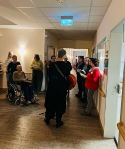 Mehrere Menschen stehen im Flur, ein Mann sitzt im Rollstuhl und eine Frau spielt auf einer Gitarre