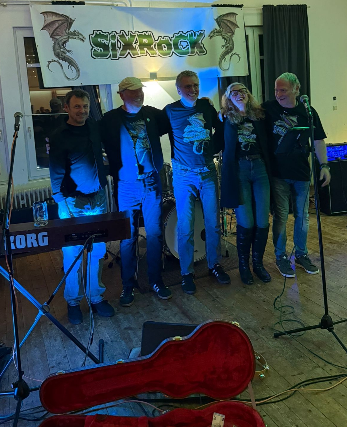 Band mit 5 Musikern stehen zwischen ihren Musikinstrumenten und unter einem Plakat mit ihrem Bandnamen Sixrock