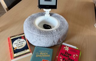 3 Bücher und ein E-Book-Reader in einer Halterung liegen bzw. stehen auf einem Tisch