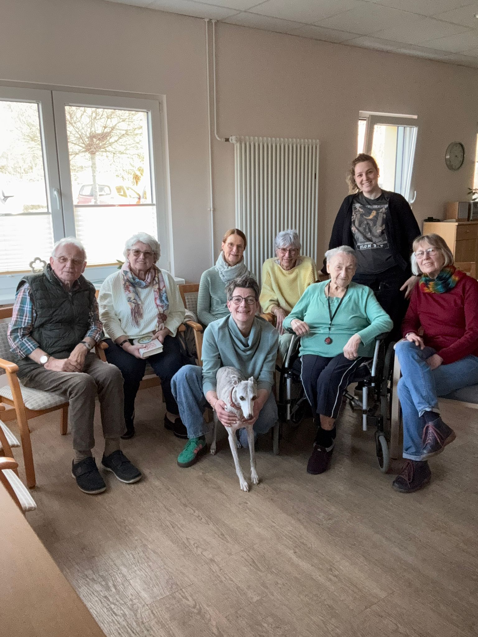 2 ehrenamtliche Mitarbeiter, ein Gast, ein Hund, ein Mann und 4 Frauen stehen oder sitzen vor einem Fenster