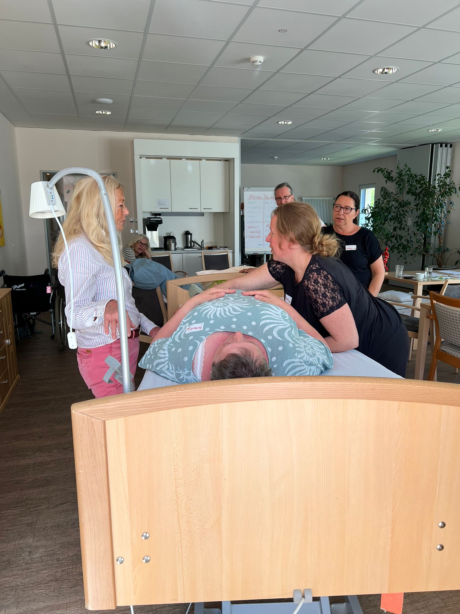 Schwester Melanie liegt in einem Bett, Schwester Sabrina greift um sie herum, Schwester Silke, Schwester Andrea und Pfleger Carsten schauen im Hintergrund zu. Die Kursleiterin erklärt etwas. Schwester Melanie liegt in einem Bett, Schwester Sabrina greift um sie herum, Schwester Silke, Schwester Andrea und Pfleger Carsten schauen im Hintergrund zu. Die Kursleiterin erklärt etwas.