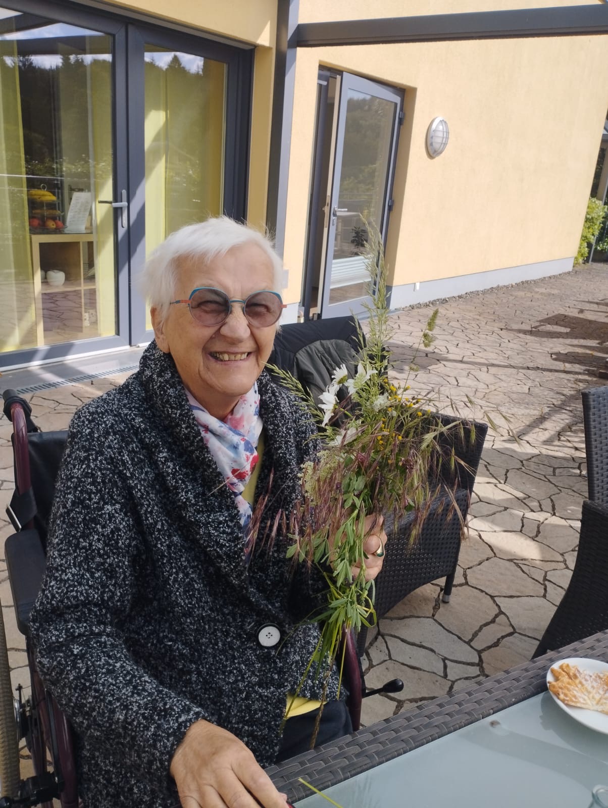 Gast sitzt im Rollstuhl auf der Terrasse und hält einen selbst gepflückten Blumenstrauß in der Hand