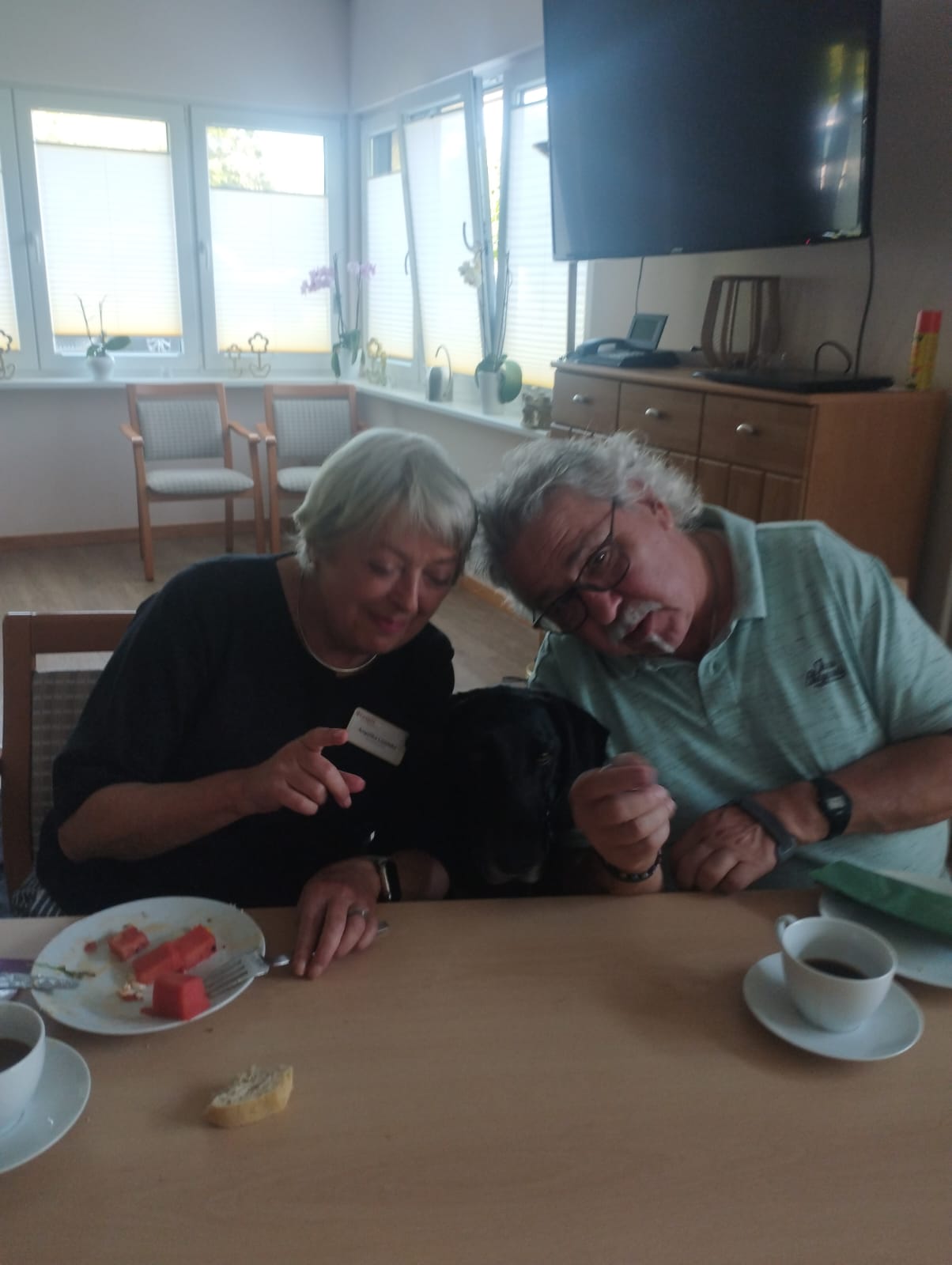 Ehrenamtliche Mitarbeiterin Dr. Angelika Lüddecke und ehrenamtlicher Hausmeister Dietmar Froböse sitzen am Tisch beim Ehrenamtstreffen. Zwischen Ihnen steht Hospiz-Hund Dako Ehrenamtliche Mitarbeiterin Dr. Angelika Lüddecke und ehrenamtlicher Hausmeister Dietmar Froböse sitzen am Tisch beim Ehrenamtstreffen. Zwischen Ihnen steht Hospiz-Hund Dako
