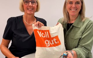 Geschäftsführerin Britta Bötel und Mitarbeiterin von der Volksbank BRAWO Salzgitter stehen an einem Tisch. Auf dem Tisch liegen Kronkorken