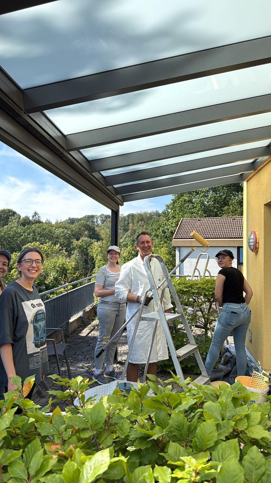 Mitarbeiter von PowerCo stehen auf der Gast-Terrasse und ein Mitarbeiter hält eine Farbrolle in der Hand Mitarbeiter von PowerCo stehen auf der Gast-Terrasse und ein Mitarbeiter hält eine Farbrolle in der Hand